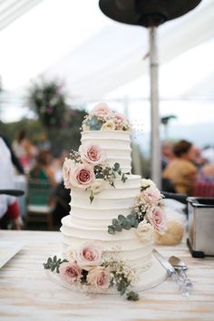 a white wedding cake with pink flowers and greenery on the top is sitting on a table