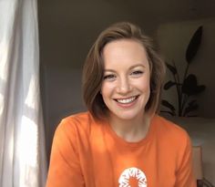 a smiling woman wearing an orange shirt in front of a window with white drapes