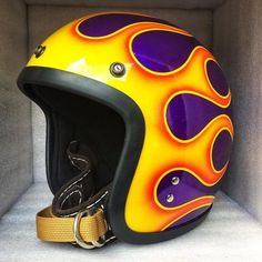 a yellow and purple helmet with flames on it sitting in a white box next to a pair of scissors