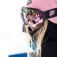 a woman wearing ski goggles and a pink hat is reflected in the mirror of her snowboarder's goggles