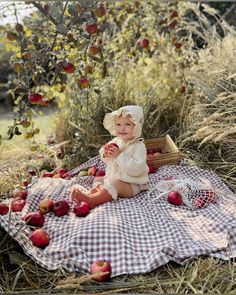 a baby sitting on a blanket in an apple orchard with apples scattered around her and wearing a bonnet