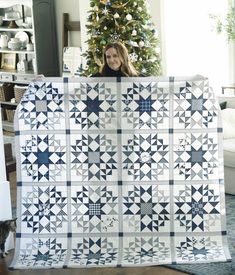 a woman holding up a blue and white quilt in front of a decorated christmas tree