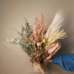 a person holding a bunch of dried flowers in their left hand, with the background gray wall behind them