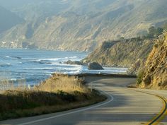 an empty road near the ocean with mountains in the background and waves crashing on the shore