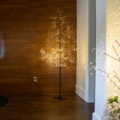 a lighted tree in the corner of a room next to a vase with flowers on it