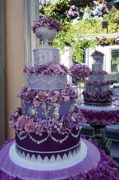 a purple wedding cake with lots of flowers on the top and bottom tier, sitting in front of a window