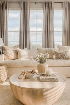 a living room with white couches and pillows on top of the coffee table in front of two windows