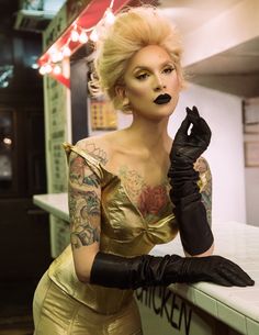 a woman in gold dress and black gloves with tattoos on her arm, leaning against a counter