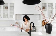 a woman is standing in the kitchen with flowers on the counter and behind her are two black vases