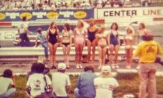 several women in bathing suits standing on the side of a race track while others watch