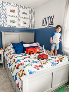 a young boy standing on top of a bed next to a toy train and truck