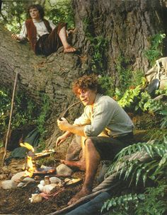 two people sitting on the ground in front of a tree and some logs with fire