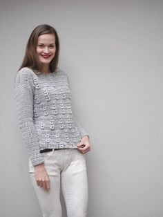 a woman standing in front of a gray wall wearing white pants and a grey sweater