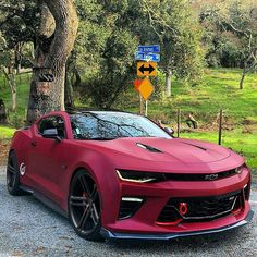 a red chevrolet camaro parked in front of a tree and street sign on the side of the road