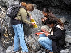 three women are sitting on the rocks and looking at their cell phones while one woman is holding a book