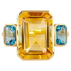 This 18 Karat Gold 7.79 Carat Blue & Yellow Topaz ""Three Stone"" Ring embodies an exquisite symphony of elegance and vibrancy. It’s a splendid testament to the seamless fusion of the serene blue and radiant yellow topaz stones. Crafted meticulously to exude luxury, this ring is a work of art that captures attention with its mesmerizing design. The ring showcases three resplendent topaz stones—a central enchanting blue topaz, flanked by two vivid yellow topaz gemstones. The striking contrast bet Luxury Three Stone Topaz Ring, Luxury Yellow Topaz Wedding Ring, Luxury Yellow Topaz Engagement Ring, Luxury Gold Citrine Topaz Ring, Luxury Yellow Gold Ring With Yellow Sapphire, Luxury Yellow Topaz Ring With Polished Finish, Luxury Yellow Topaz Ring With Yellow Sapphire, Luxury Yellow Sapphire Jewelry In Emerald Cut, Luxury Yellow Topaz Ring With Prong Setting