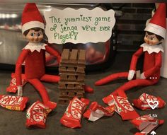 two elfs are sitting next to a pile of candy and one is holding a sign