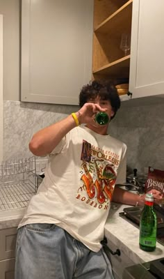 a man standing in a kitchen drinking from a green bottle with his hand on the counter