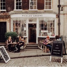 some people sitting at tables in front of a building with windows and signs on the sidewalk