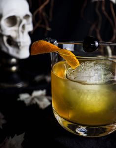 a halloween cocktail with an orange peel garnished in ice and a skull behind it