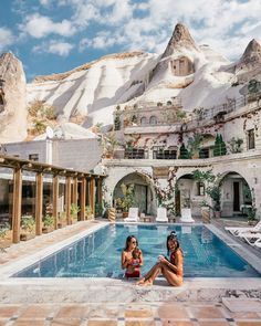 a woman sitting on the edge of a swimming pool in front of a mountain range