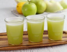 three glasses filled with green liquid on top of a wooden tray next to some fruit