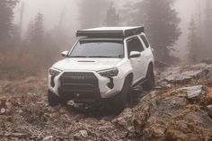 a white four - doored vehicle driving on rocks in the woods with fog behind it