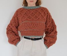 a woman standing in front of a white wall wearing an orange sweater and beige pants