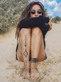 a person sitting in the sand with tattoos on their legs and feet, wearing sunglasses