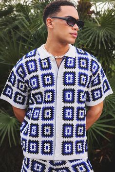 a man wearing sunglasses and a crocheted shirt stands in front of palm trees