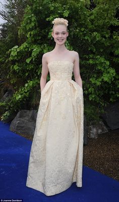 a woman in a white dress standing on a blue carpet with trees behind her and bushes to the side