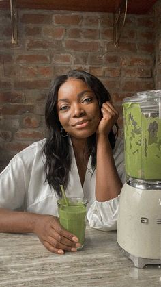 a woman sitting at a table next to a blender filled with green smoothie