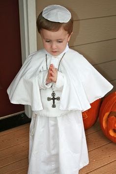 a little boy dressed in a priest outfit and holding his hands together while standing next to a pumpkin