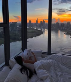 a woman is laying in bed looking out the window at the river and cityscape