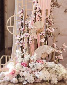 an arrangement of flowers and fan on a table in front of a wall with pink drapes