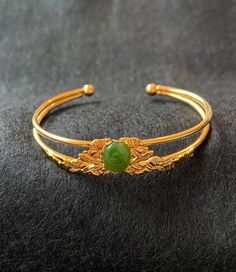 Vintage gold plated open bangle cuff bracelet with BC nephrite jade cabochon. Nephrite cabochon is 8 mm by 10 mm. The cabochon is mounted on a gold leaf and ribbon textural design. Bracelet arms are 3 mm thick. The balls at the inside ends of the cuff bracelet are 5 mm thick.  The bracelet is 60 mm thick. Nephrite jade is recognizable by its dark olive green, lightly translucent, colour. This jade was mined in British Columbia in the mid-1960s. Gold Cabochon Bangle Jewelry, Gold Jade Bangle Bracelet, Elegant Gold Jade Bracelets, Gold Jade Bangle, Handmade Elegant Jade Bangle, Luxury Gold Jade Bracelets, Formal Gold Jade Bracelets, Design Bracelet, Jade Bangle