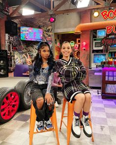 two young women sitting on stools in front of a tv set with an orange car behind them