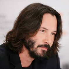 a man with long hair and beard wearing a black suit sitting in front of a white wall