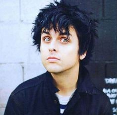 a young man with black hair and piercings looks into the distance while standing in front of a brick wall