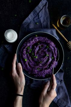 two hands holding a bowl of purple mashed potatoes on a black table with spoons