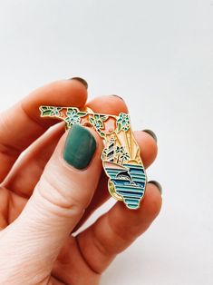 a woman's hand holding a green and gold pin with a lake scene on it