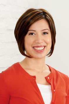 a smiling woman in an orange cardigan smiles at the camera while standing next to a white brick wall