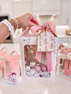 a person is holding a small gift box with pink ribbon and ornaments in it on a kitchen counter