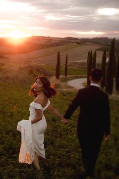 a woman in a white dress and a man in a black suit are walking through the grass