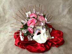 a white mask with pink flowers and sticks sticking out of it sitting on a red cloth