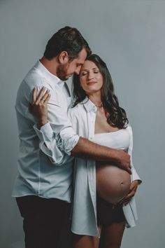 a pregnant couple cuddles and kisses while posing for a photo in front of a gray background