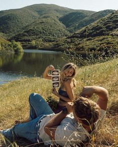 a man and woman sitting on the ground next to a lake with a bottle in their hand