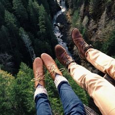 two people wearing brown shoes standing on a wooden bridge over a river in the woods