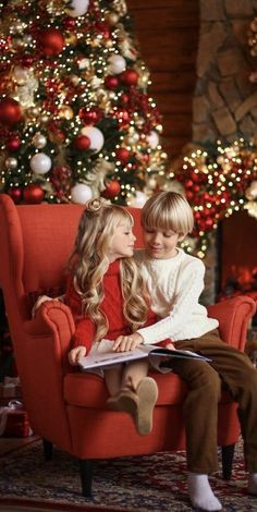 two children sitting on a red chair in front of a christmas tree