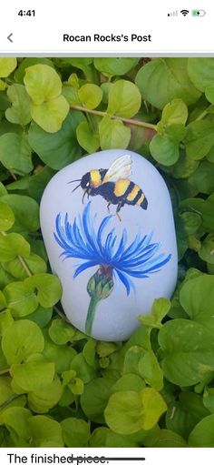 a painted rock with a bee on it sitting in some green leaves next to plants